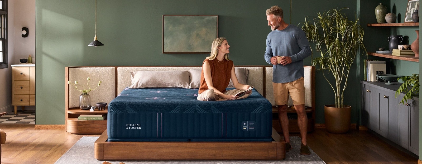 med/sm styled room with woman sitting on legacy mattress and man standing beside