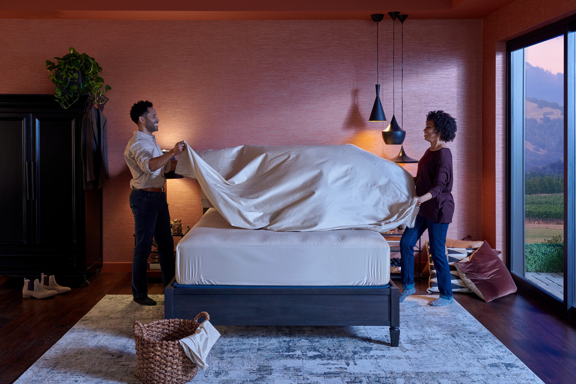 couple making their bed in a mauve colored room with 3 black lamps hanging abov ethe night stand
